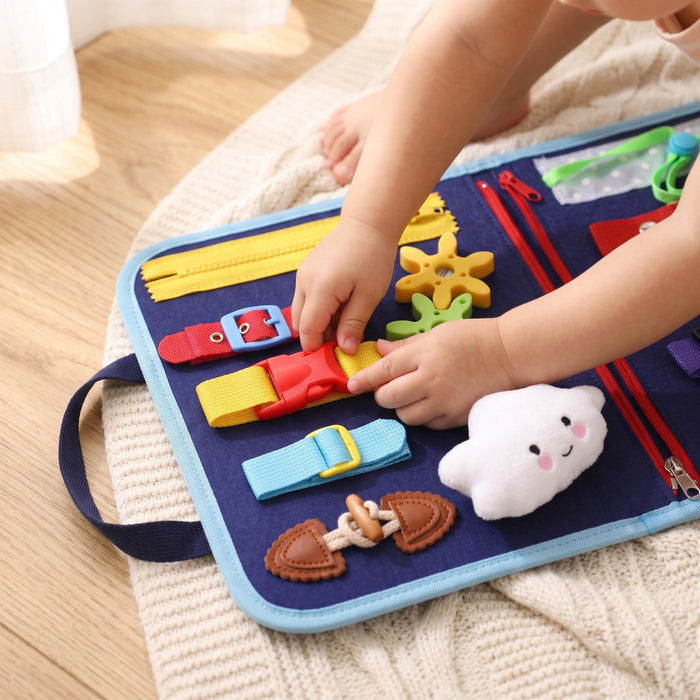 A toddler's hands press the red buckle on a yellow strap, on the open blue board lying on a knit blanket