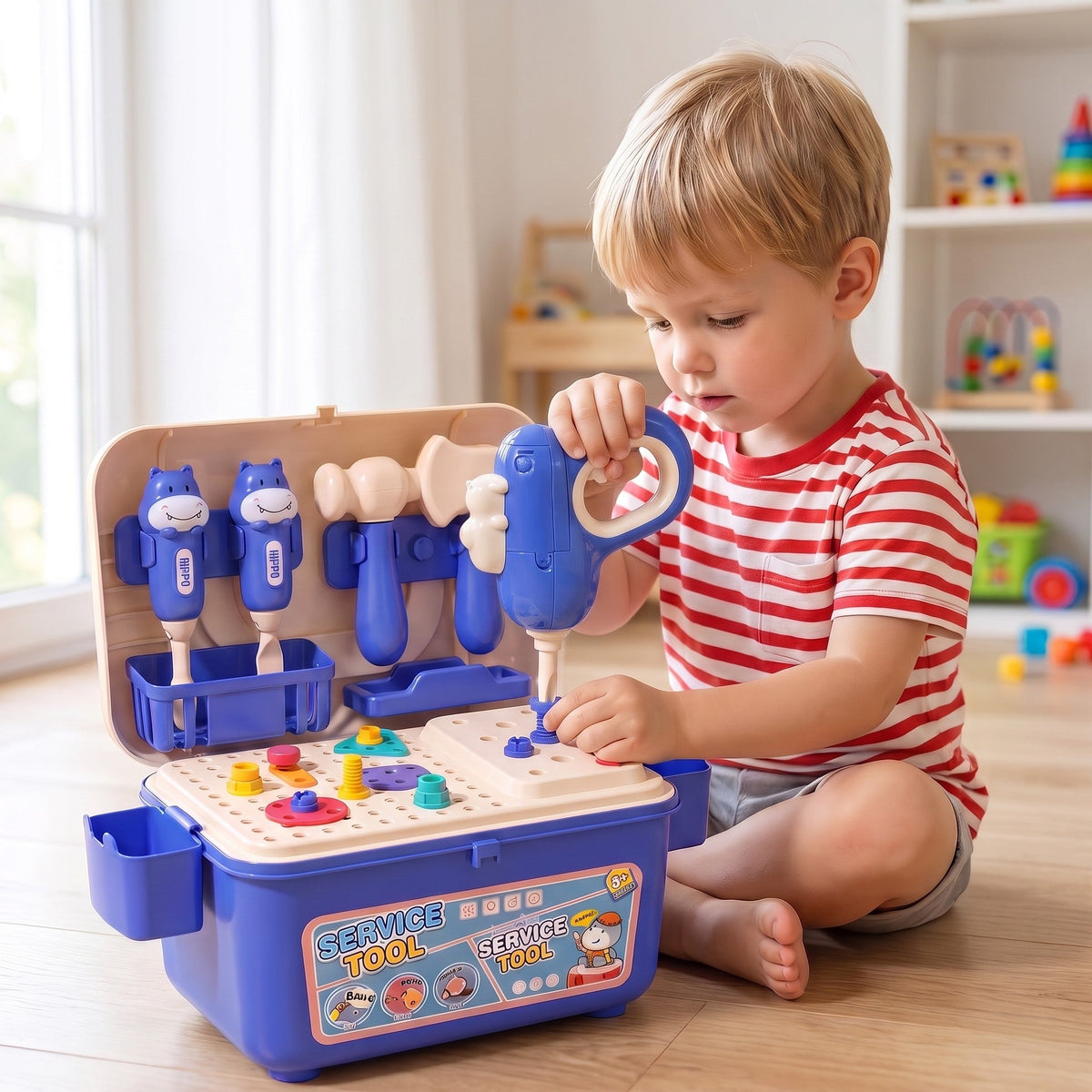 A child on the floor uses the toy drill on the toolbox pegboard, with screwdrivers, a hammer and an axe in the open lid