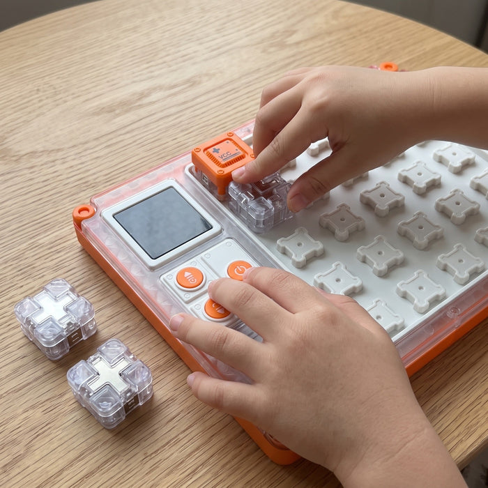 Hands snap a clear block onto the circuit board beside the orange plus block and press a button, with two spare blocks nearby