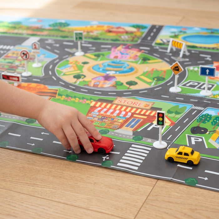 A child's hand pushes a red toy car along a road on the mat, past a traffic light, a yellow taxi and small road signs