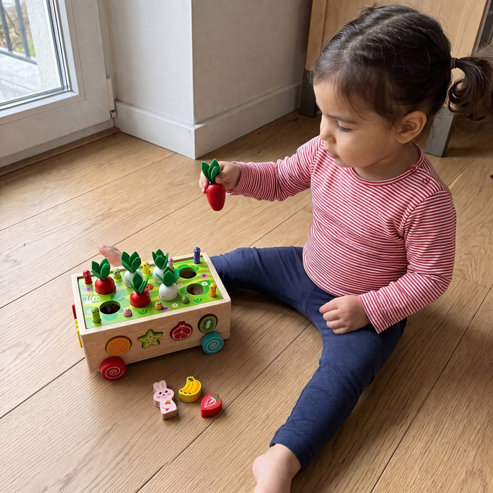 A toddler sits on the floor holding a red radish beside the wooden garden sorter, with the rabbit and fruit blocks nearby