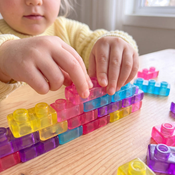 Close-up of a toddler's hands pressing a pink block onto a low wall of see-through coloured blocks on a table