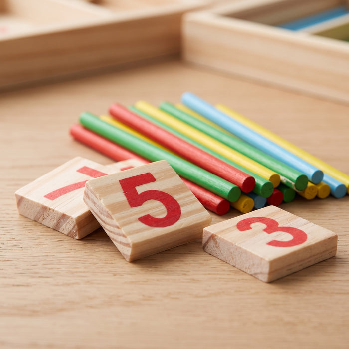 Wooden number tiles 7, 5 and 3 up close, with a pile of red, green, yellow and blue counting sticks behind them.