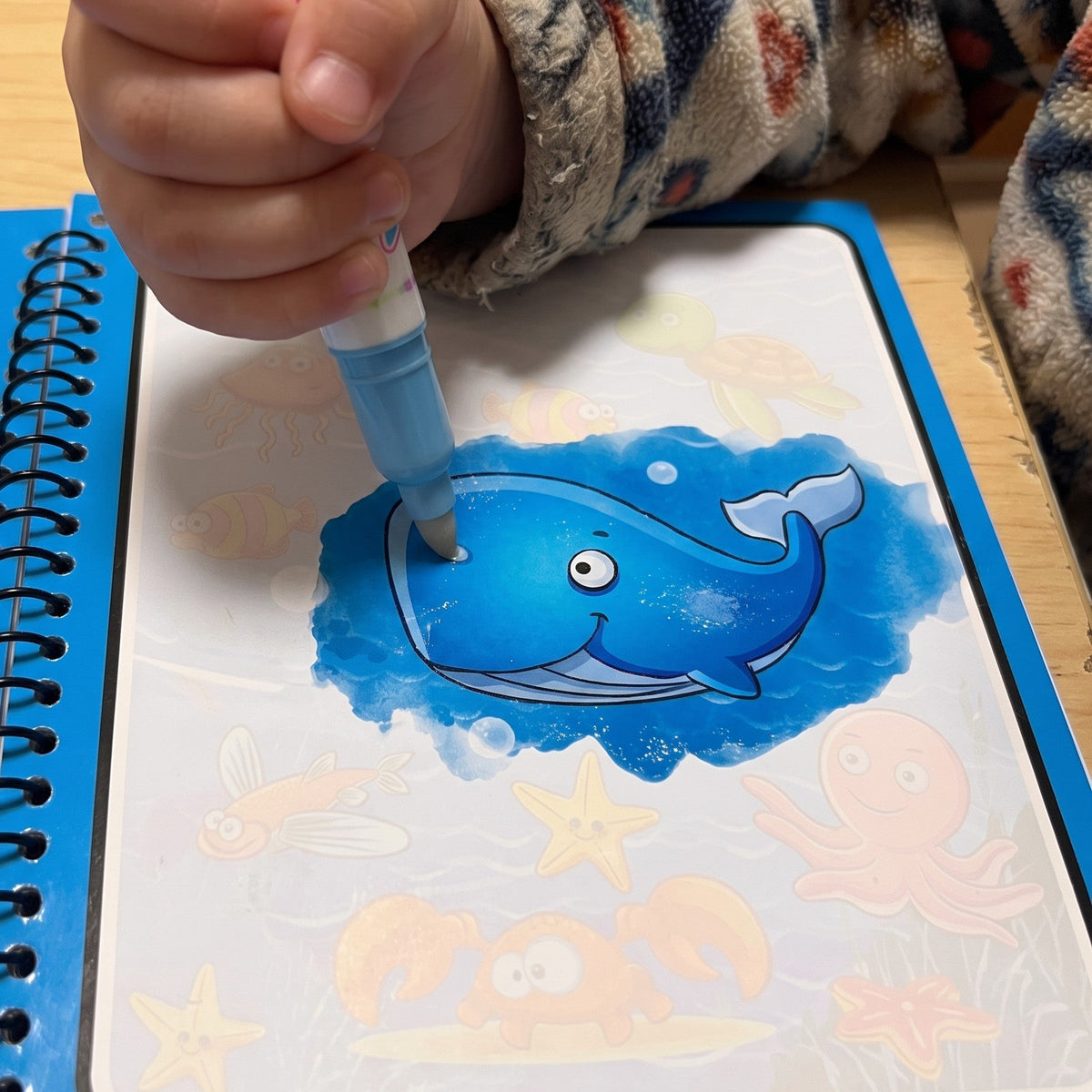 A child runs a water pen over a pale page, and a bright blue whale shows where it is wet, with faint sea animals around it