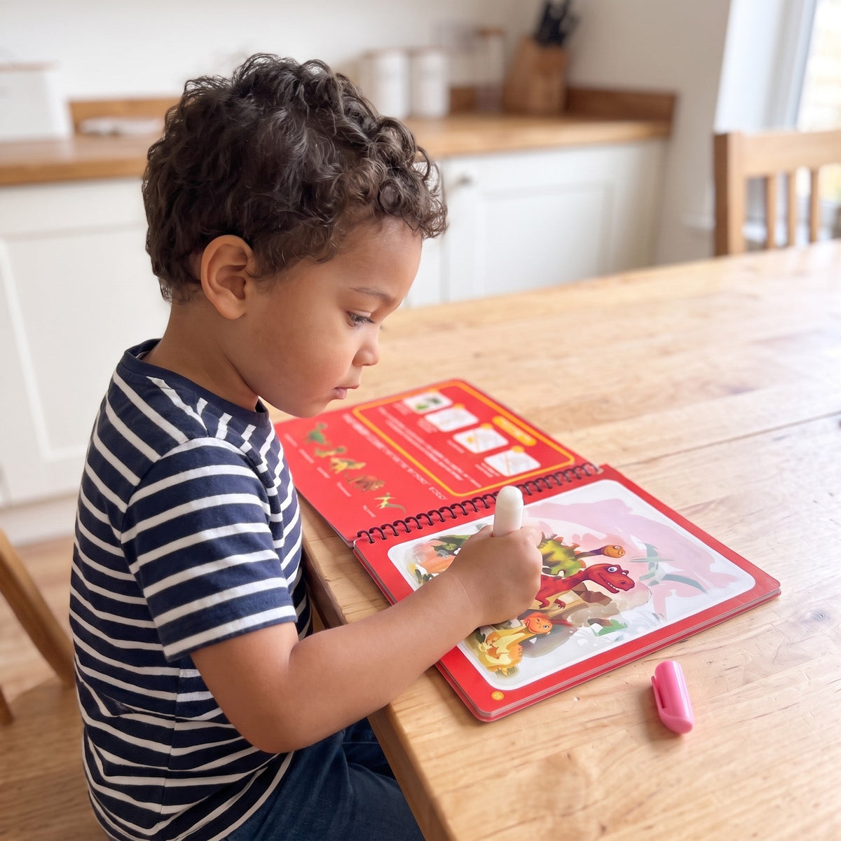 A child at a kitchen table uses a water pen on an open page of the red dinosaur book, with the pen's pink cap on the table